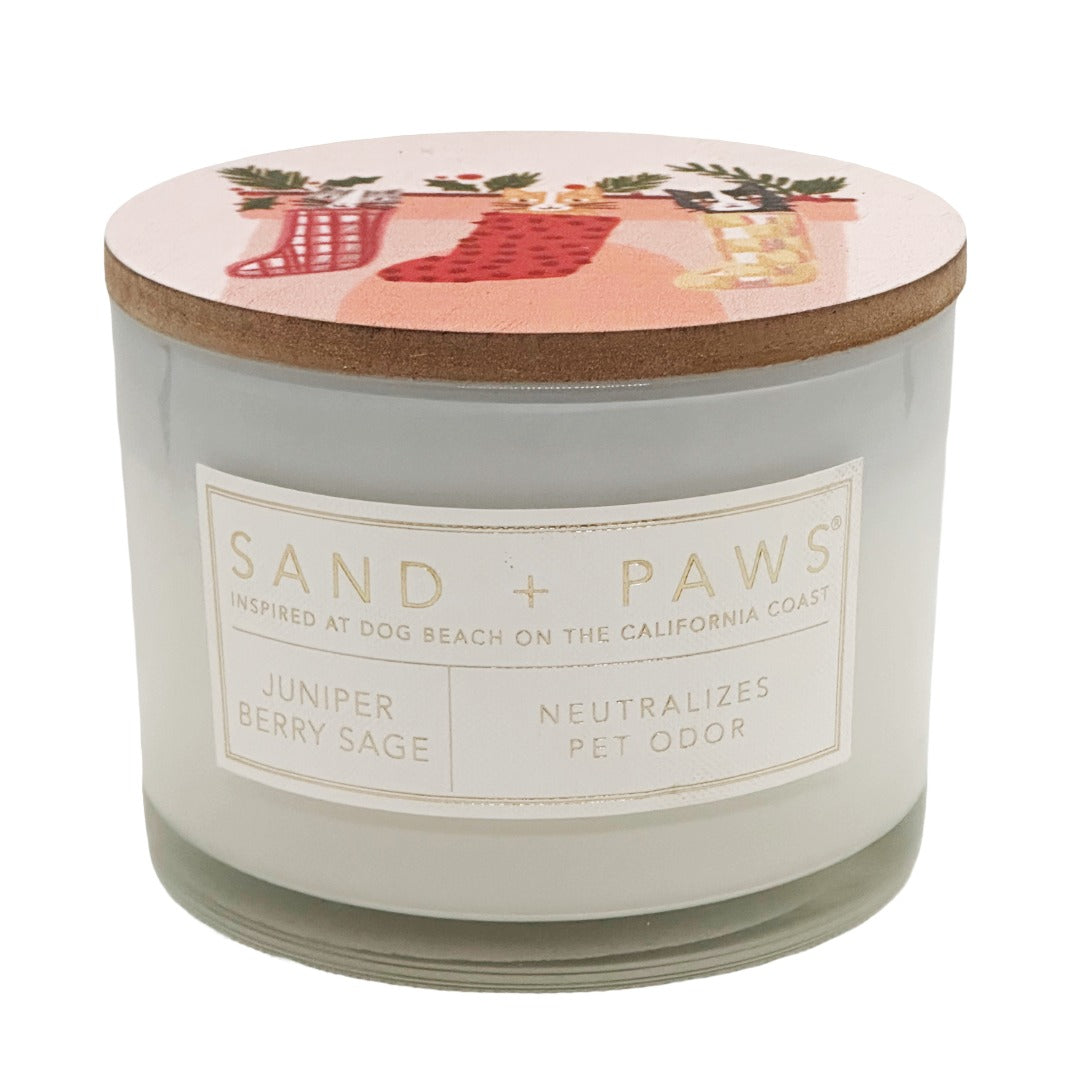 A jar candle from Sand + Paws with a transparent glass body, a cork lid, and a white label reading 'SAND + PAWS' and 'Juniper Berry Sage'. The lid has a pink holiday illustration with red stockings and greenery.
