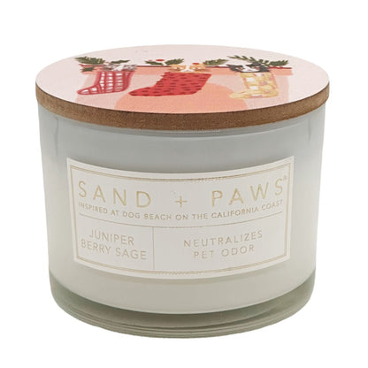 A jar candle from Sand + Paws with a transparent glass body, a cork lid, and a white label reading 'SAND + PAWS' and 'Juniper Berry Sage'. The lid has a pink holiday illustration with red stockings and greenery.