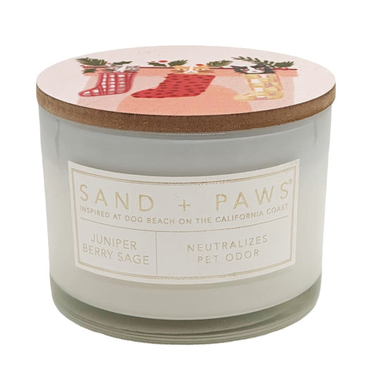 A jar candle from Sand + Paws with a transparent glass body, a cork lid, and a white label reading 'SAND + PAWS' and 'Juniper Berry Sage'. The lid has a pink holiday illustration with red stockings and greenery.