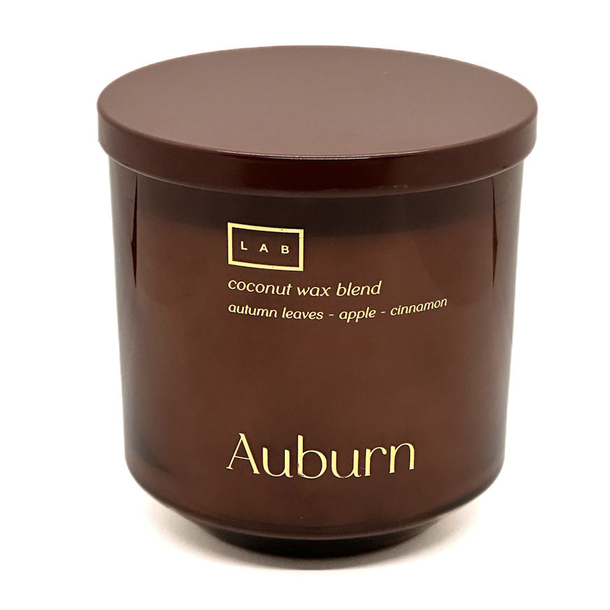 A dark amber-brown glass jar candle with a matching lid, featuring gold lettering that reads 'Auburn' and 'coconut wax blend' on a clean white background.