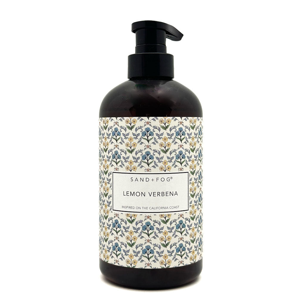 A dark amber glass pump bottle of Sand + Fog Lemon Verbena hand wash, featuring a black pump and a decorative label with a blue, green, and gold botanical pattern on a white background.