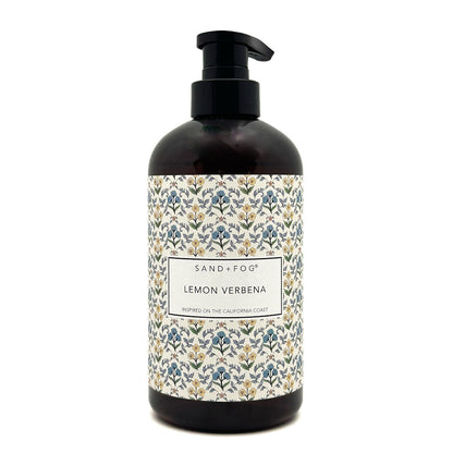 A dark amber glass pump bottle of Sand + Fog Lemon Verbena hand wash, featuring a black pump and a decorative label with a blue, green, and gold botanical pattern on a white background.