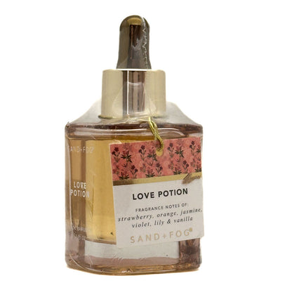 A bottle of Sand + Fog Love Potion perfume oil with a dropper cap, featuring amber liquid, a pink floral label, and a white text label on a white background.