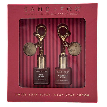 Sand + Fog perfume oil gift set in a burgundy box, containing Love Potion Perfume Oil and Strawberry Spritz Perfume  Oil, each with a gold keychain and round medallion.