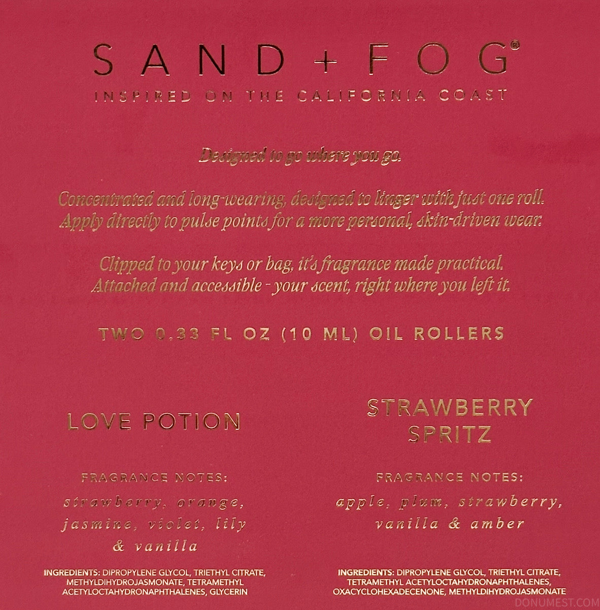 A close-up of a Sand + Fog fragrance oil card showing the 'Love Potion' and 'Strawberry Spritz' scents on a deep red background with gold text.
