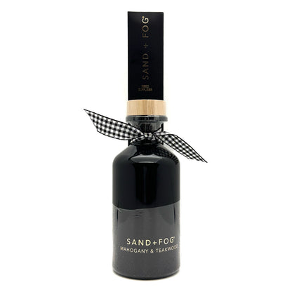 A dark glass bottle of Sand + Fog Mahogany & Teakwood fragrance with a black cap, a light wood collar, and a black-and-white gingham ribbon tied around the neck, on a white background.