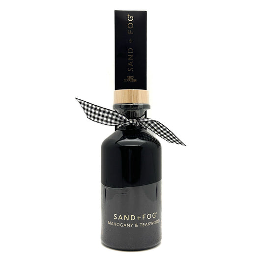 A dark glass bottle of Sand + Fog Mahogany & Teakwood fragrance with a black cap, a light wood collar, and a black-and-white gingham ribbon tied around the neck, on a white background.