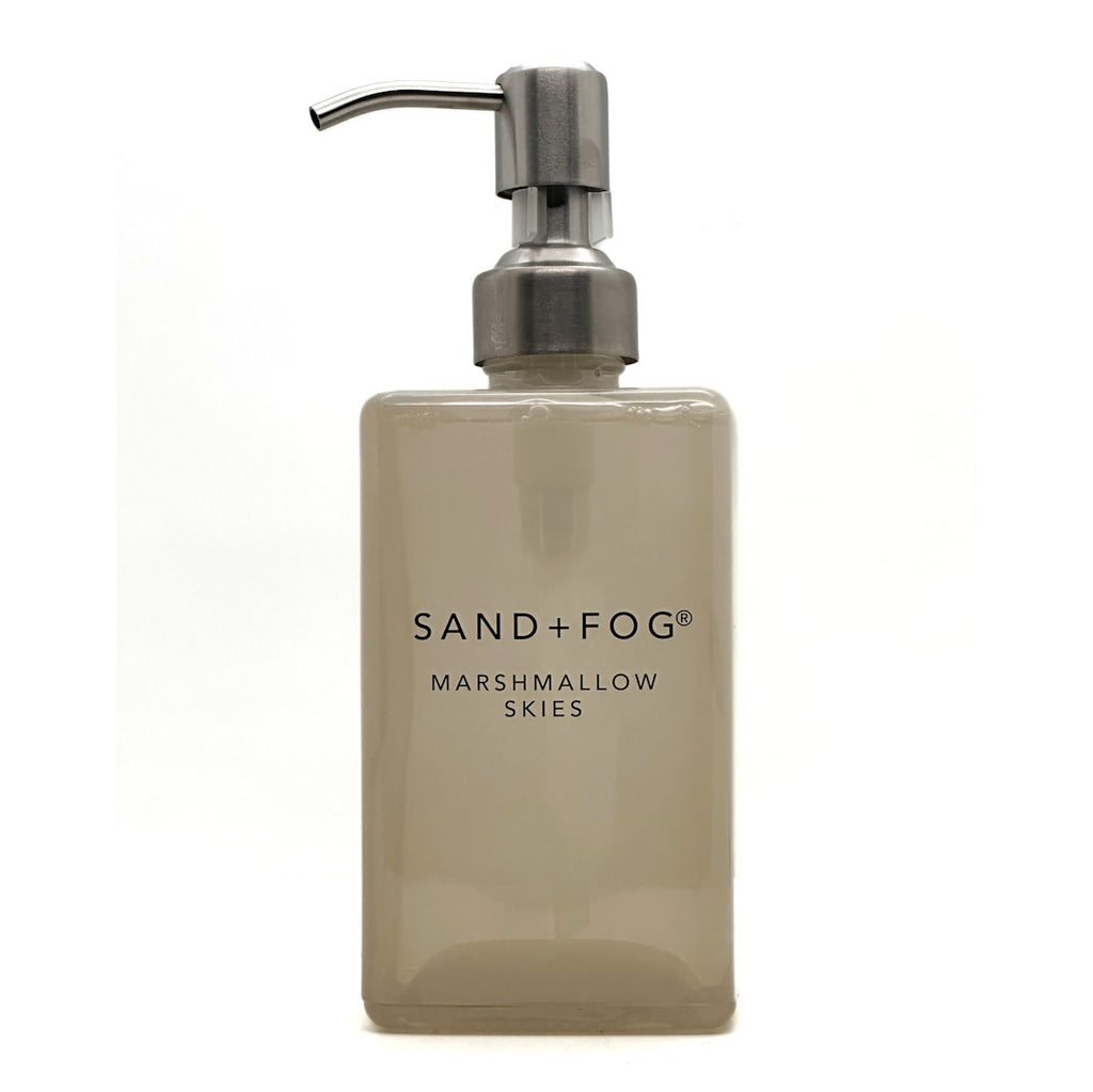 A frosted glass soap dispenser bottle in a warm beige-tinted shade with a brushed silver metal pump, featuring the text 'SAND+FOG MARSHMALLOW SKIES' on the front, set against a plain white background.