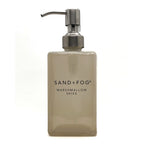 A frosted glass soap dispenser bottle in a warm beige-tinted shade with a brushed silver metal pump, featuring the text 'SAND+FOG MARSHMALLOW SKIES' on the front, set against a plain white background.