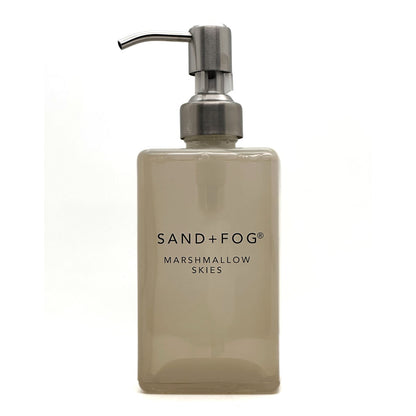 A frosted glass soap dispenser bottle in a warm beige-tinted shade with a brushed silver metal pump, featuring the text 'SAND+FOG MARSHMALLOW SKIES' on the front, set against a plain white background.