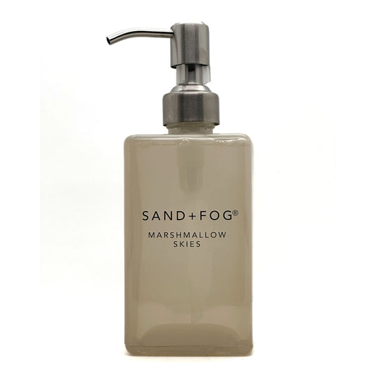 A frosted glass soap dispenser bottle in a warm beige-tinted shade with a brushed silver metal pump, featuring the text 'SAND+FOG MARSHMALLOW SKIES' on the front, set against a plain white background.