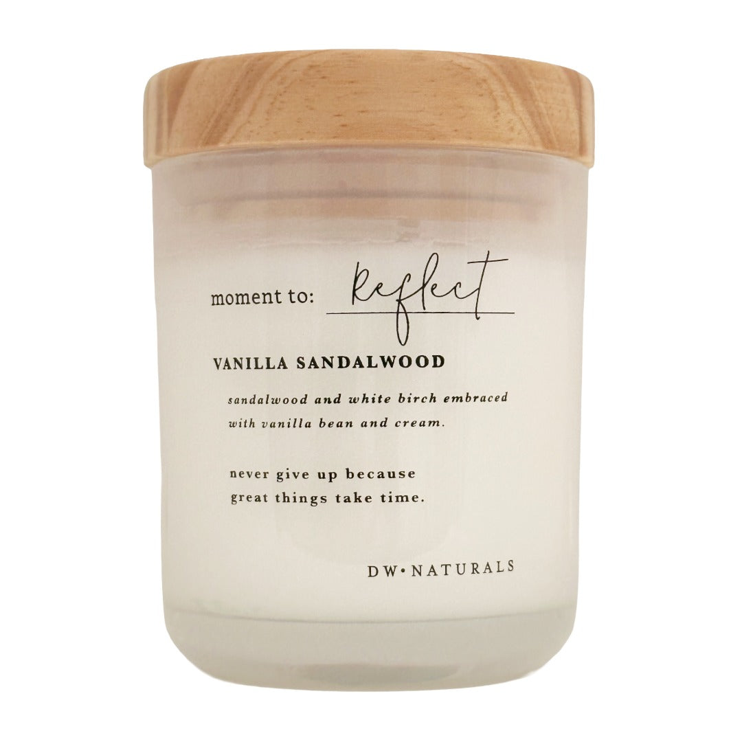 A candle in a frosted glass jar with a light wood lid, featuring black text that reads 'moment to: Reflect' and 'VANILLA SANDALWOOD' on a clean white background.