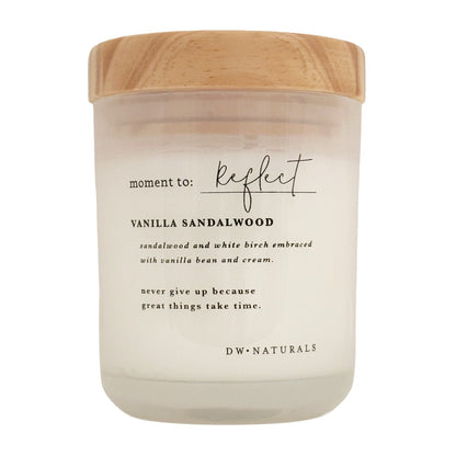 A candle in a frosted glass jar with a light wood lid, featuring black text that reads 'moment to: Reflect' and 'VANILLA SANDALWOOD' on a clean white background.