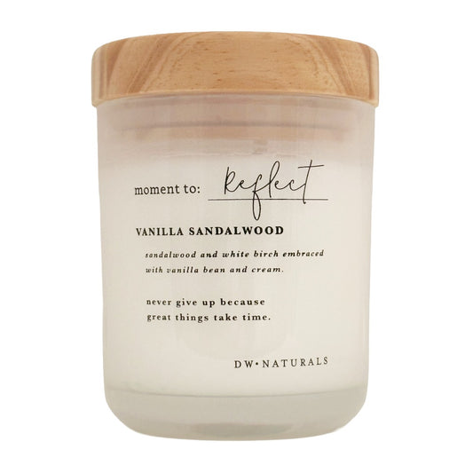 A candle in a frosted glass jar with a light wood lid, featuring black text that reads 'moment to: Reflect' and 'VANILLA SANDALWOOD' on a clean white background.