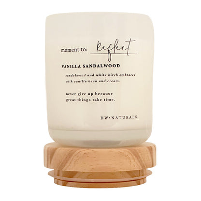 A candle in a frosted cream glass jar with a light natural wood base, labeled 'moment to: reflect' and 'VANILLA SANDALWOOD' by DW NATURALS, on a clean white background.
