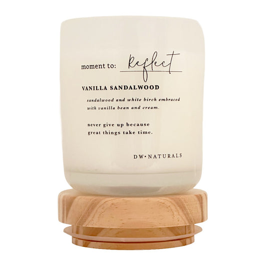 A candle in a frosted cream glass jar with a light natural wood base, labeled 'moment to: reflect' and 'VANILLA SANDALWOOD' by DW NATURALS, on a clean white background.