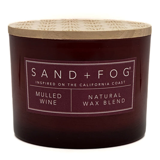 A dark red glass jar candle with a light wood lid, featuring a label that reads 'SAND+FOG' and 'MULLED WINE NATURAL WAX BLEND'.