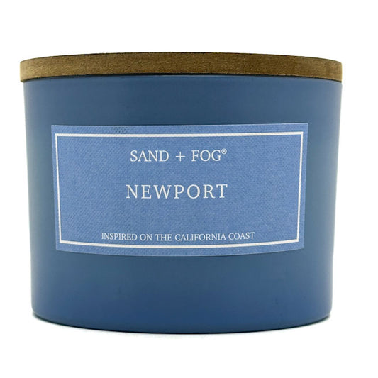 Blue candle with a wooden lid labeled 'Sand + Fog Newport' on a white background