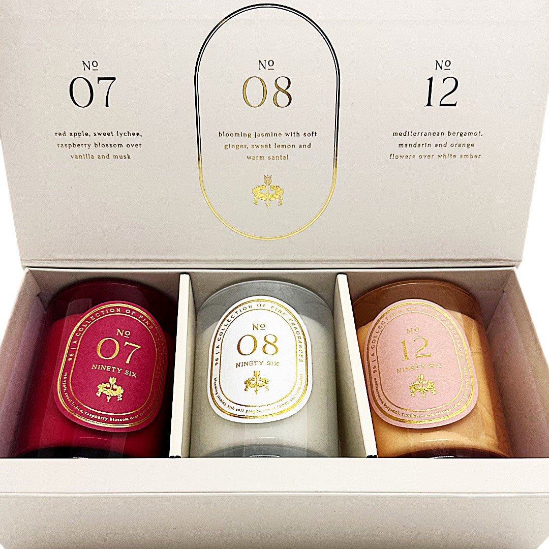An open Ninety Six candle gift set box showing three candles in different colours: red, pale green, and peach, with printed labels and scent names on the inside lid.