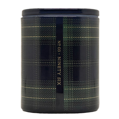 A cylindrical candle with a dark navy and green tartan plaid pattern, featuring a vertical black band with gold text reading 'Nº 03 · Ninety Six'.