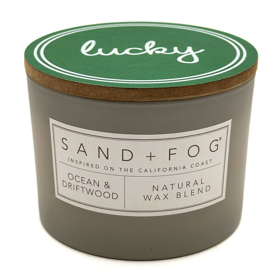 Candle with 'lucky' lid, Sand + Fog brand label, and 'Ocean & Driftwood' description on a white background