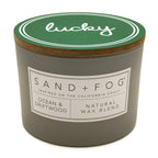 Candle with 'lucky' lid, Sand + Fog brand label, and 'Ocean & Driftwood' description on a white background
