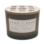 Candle with 'Sand + Paws' label and ocean and driftwood scent on a white background