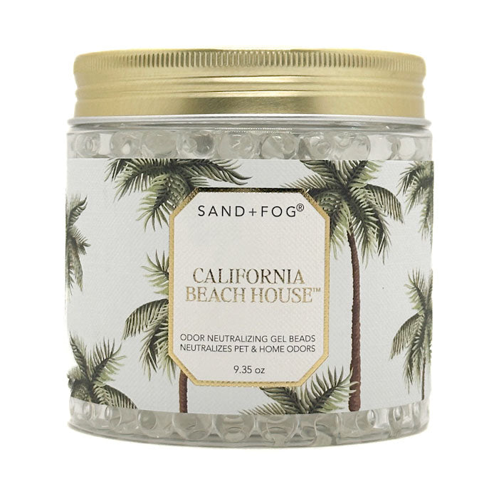 A clear plastic jar filled with gel beads, featuring a gold metal lid and a white label with a gold border and green palm tree illustrations. The label reads 'SAND+FOG' and 'CALIFORNIA BEACH HOUSE'.