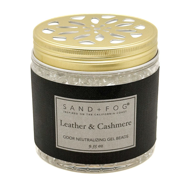 A jar of Sand + Fog Leather & Cashmere odor neutralizing gel beads, featuring a black textured label and a gold metal lid with a decorative cutout pattern, set against a plain white background.