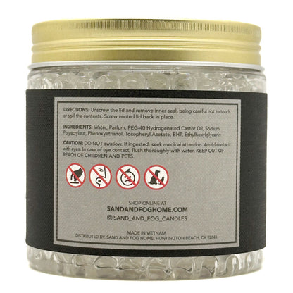 A clear plastic jar filled with white beads, featuring a gold screw-on lid and a large black label with product instructions, warning icons, and the Sand and Fog Candle brand name.