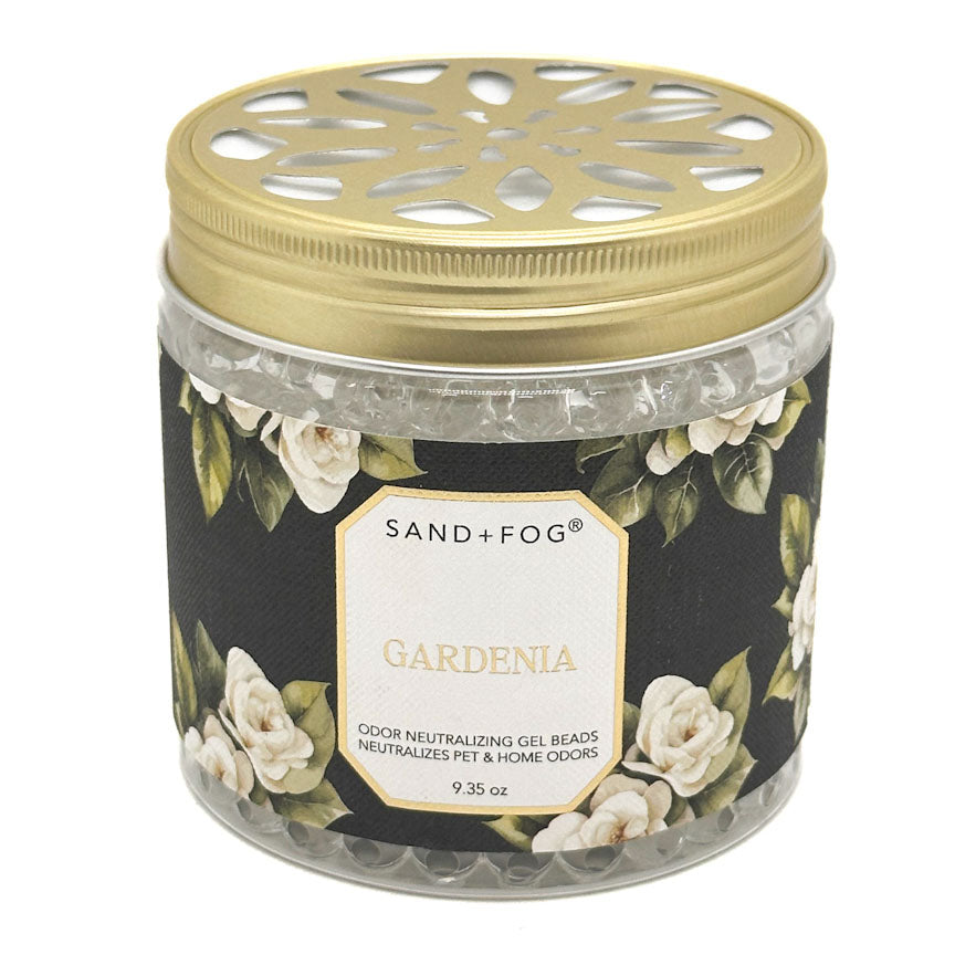 A Sand + Fog Gardenia odour neutralising gel beads candle in a clear glass jar with a gold metal lid and a black floral label featuring white roses.