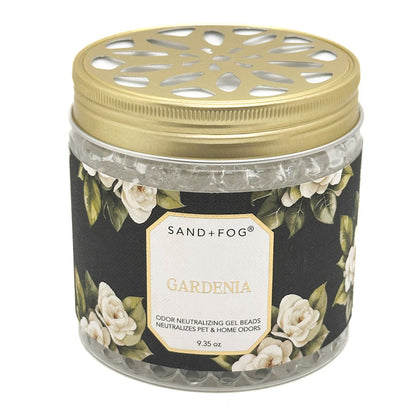 A Sand + Fog Gardenia odour neutralising gel beads candle in a clear glass jar with a gold metal lid and a black floral label featuring white roses.