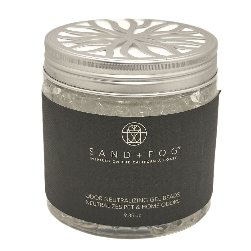 A clear plastic jar filled with clear teakwood gel beads, featuring a silver metal lid with a decorative cutout pattern and a black paper label. The label displays the brand 'SAND + FOG' in white text.