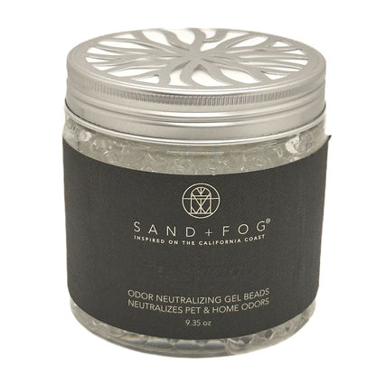 A clear plastic jar filled with clear teakwood gel beads, featuring a silver metal lid with a decorative cutout pattern and a black paper label. The label displays the brand 'SAND + FOG' in white text.