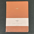 An Osprey London lined notebook in a peachy orange textured leather cover, wrapped in clear plastic and featuring a white paper band label around the middle.