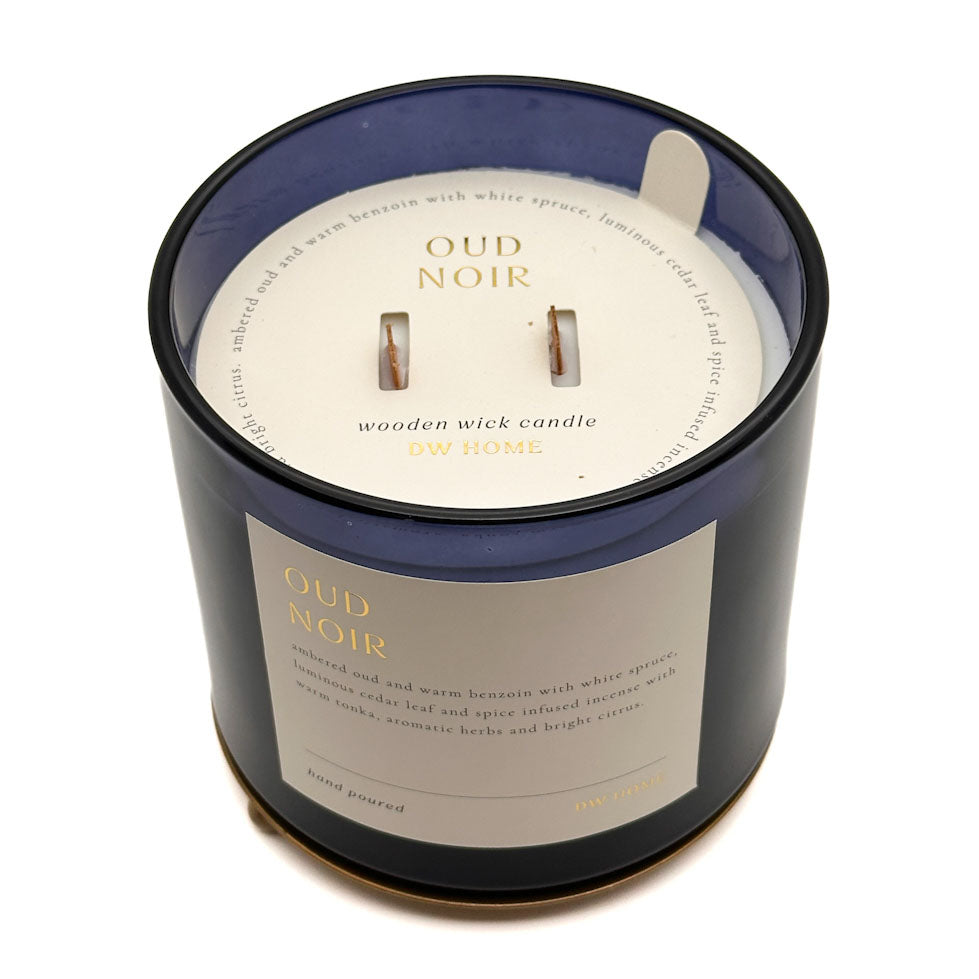 A dark blue glass jar candle with a cream-coloured label and wick insert, photographed on a clean white background. The label reads 'OUD NOIR' and 'wooden wick candle ON HOME'.