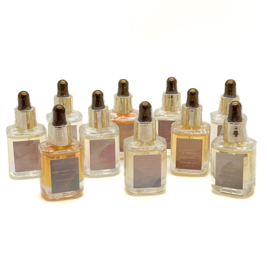A SAND + FOG collection of small clear glass perfume oil dropper bottles with metallic caps and square labels, arranged on a plain white background. The bottles contain liquids in warm amber, pink, and brown tones.