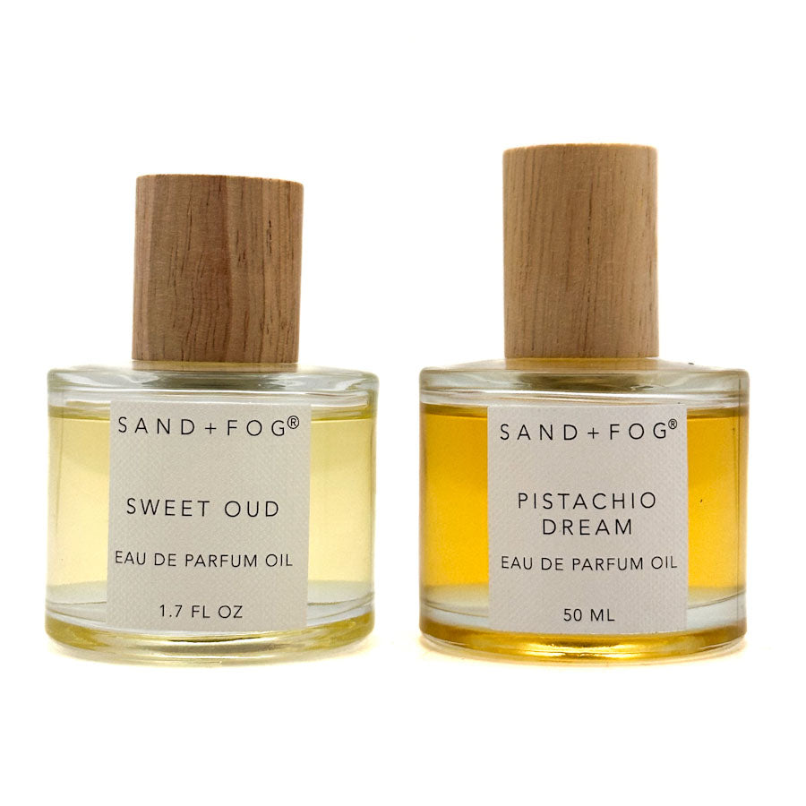 Two clear glass bottles of Sand + Fog Eau de Parfum Oil with light wood caps, shown side by side on a white background. 