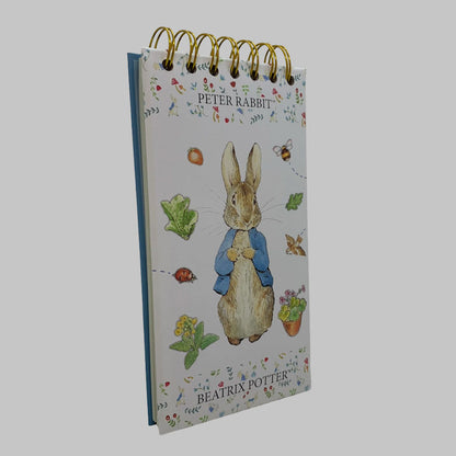A Peter Rabbit spiral-bound notebook or desk pad with gold wire binding, featuring an illustration of Peter Rabbit in a blue jacket on a white background with floral details and the text 'PETER RABBIT' and 'BEATRIX POTTER'.