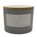 A dark grey jar candle with a light wood lid and a grey label that reads 'SAND+FOG' and 'PINEAPPLE COCONUT NATURAL WAX BLEND'.