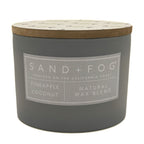 A dark grey jar candle with a light wood lid and a grey label that reads 'SAND+FOG' and 'PINEAPPLE COCONUT NATURAL WAX BLEND'.