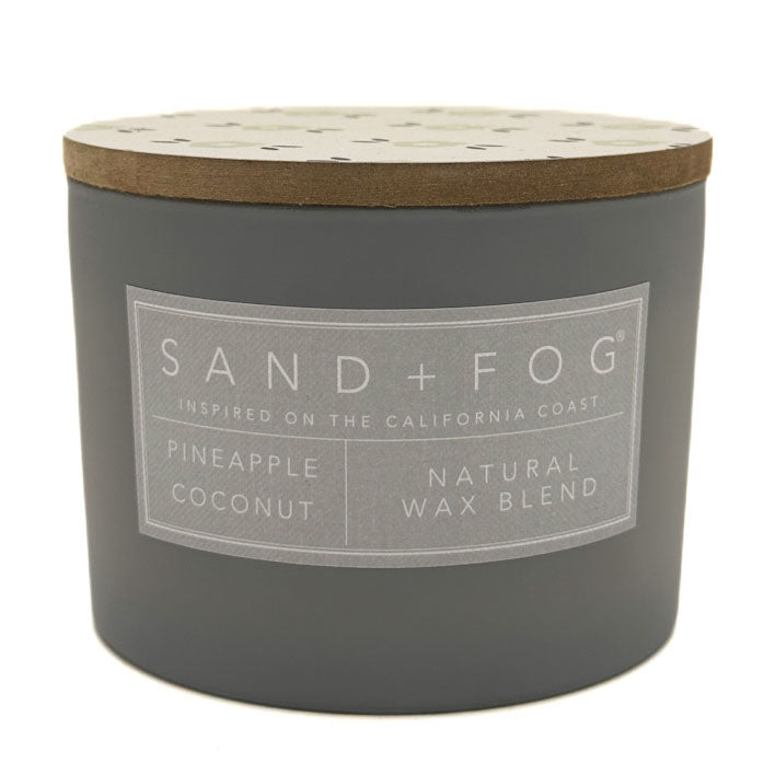 A dark grey jar candle with a light wood lid and a grey label that reads 'SAND+FOG' and 'PINEAPPLE COCONUT NATURAL WAX BLEND'.