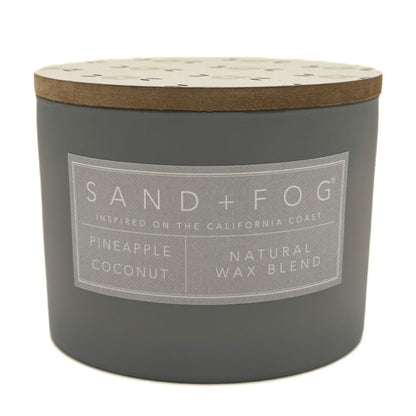 A dark grey jar candle with a light wood lid and a grey label that reads 'SAND+FOG' and 'PINEAPPLE COCONUT NATURAL WAX BLEND'.