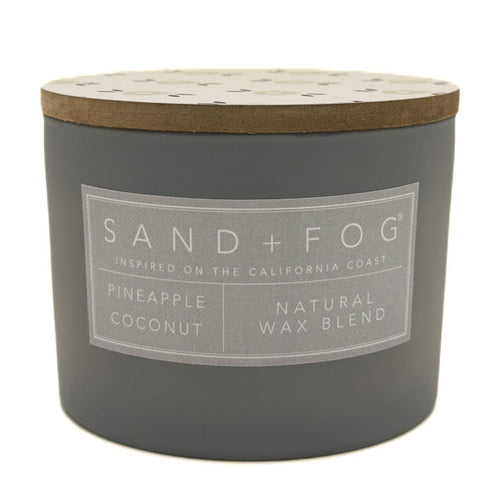 A dark grey jar candle with a light wood lid and a grey label that reads 'SAND+FOG' and 'PINEAPPLE COCONUT NATURAL WAX BLEND'.