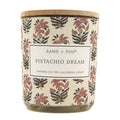A jar candle with a light wood lid and a cream-colored label featuring a pink, purple, and dark gray floral pattern. The label reads 'SAND + FOG PISTACHIO DREAM'.