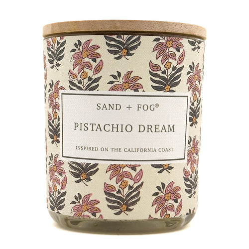 A jar candle with a light wood lid and a cream-colored label featuring a pink, purple, and dark gray floral pattern. The label reads 'SAND + FOG PISTACHIO DREAM'.