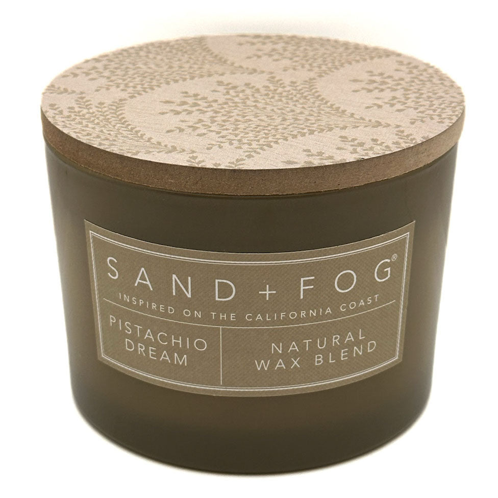 A green jar candle with a patterned beige lid and a white label reading 'SAND+FOG' and 'Pistachio Dream Natural Wax Blend'.
