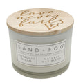 A candle jar with a white wax body, a light wood lid, and the words 'Love never fails' on lid. The front label reads SAND + FOG, PISTACHIO DREAM, and NATURAL WAX BLEND.