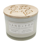 A candle jar with a white wax body, a light wood lid, and the words 'Love never fails' on lid. The front label reads SAND + FOG, PISTACHIO DREAM, and NATURAL WAX BLEND.