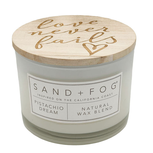 A candle jar with a white wax body, a light wood lid, and the words 'Love never fails' on lid. The front label reads SAND + FOG, PISTACHIO DREAM, and NATURAL WAX BLEND.